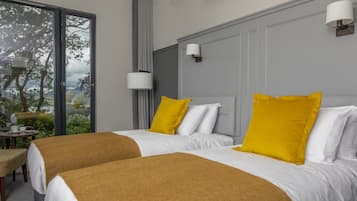 A hotel room with two beds, each with white bedding and a mustard-colored throw blanket. There are two bedside lamps, a window with a view of trees and hills, and a small table with a chair.