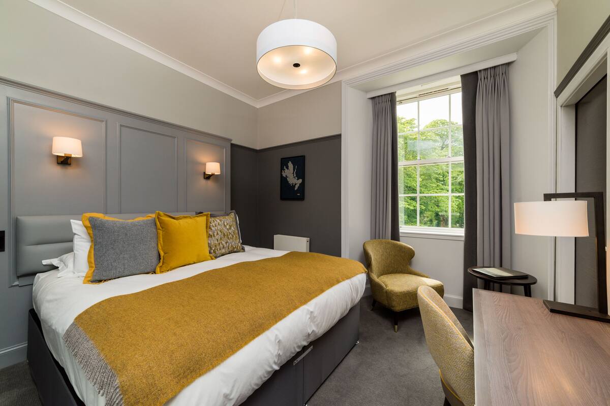 A hotel room with a large bed, a desk, a chair, a window with curtains, and wall-mounted lamps.