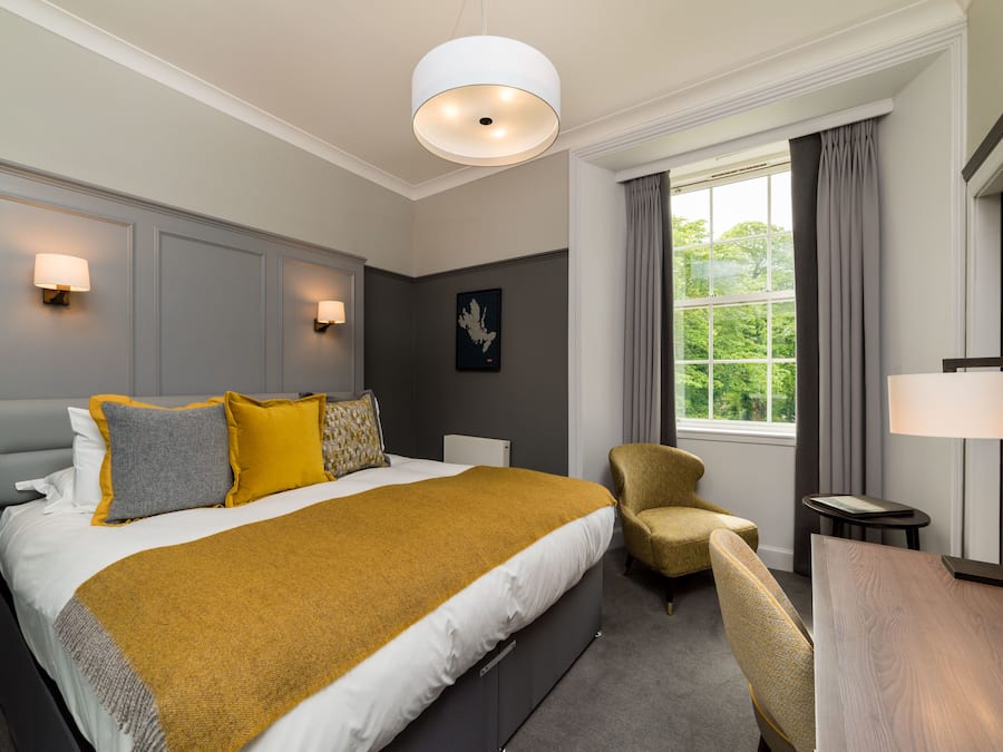 A hotel room with a large bed, a desk, a chair, a window with curtains, and wall-mounted lamps.
