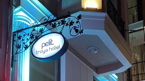 Fachada de un hotel con letreros iluminados que indican "petit troya hotel" y un mostrador de recepción visible a través de las puertas de vidrio.