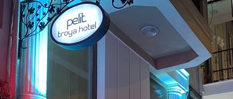A hotel entrance with illuminated signage reading "petit troya hotel" and a reception desk visible through the glass doors.
