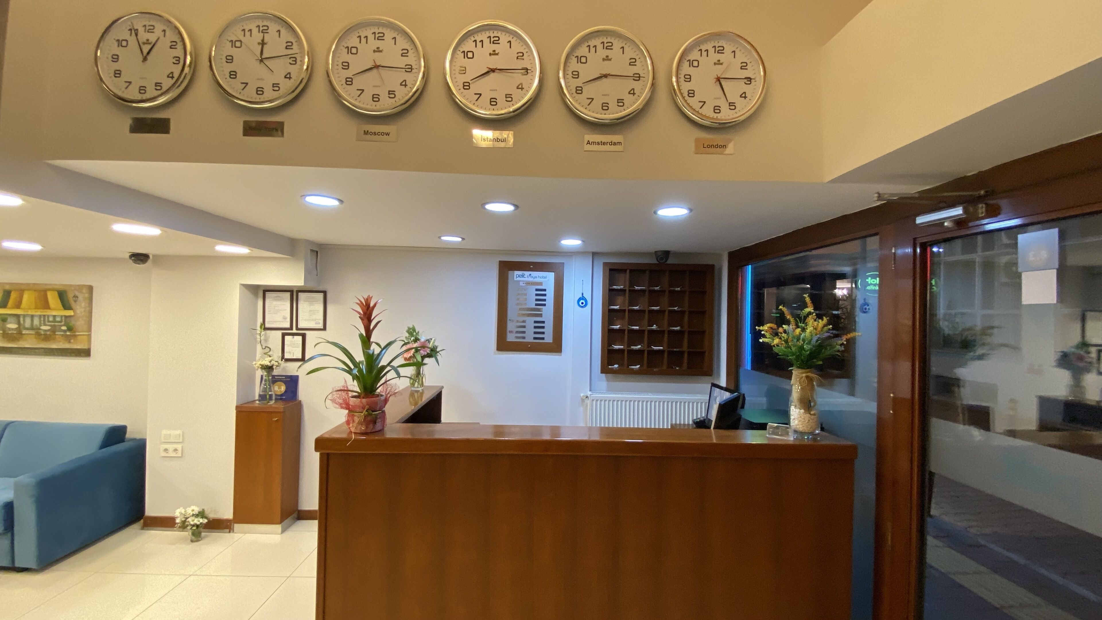 A reception area with a wooden desk, potted plants, framed certificates, and clocks indicating different cities.