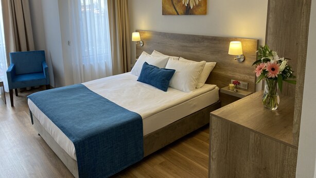 Una habitación de hotel con una cama, dos lámparas de noche, un cuadro, una silla y un florero con flores.