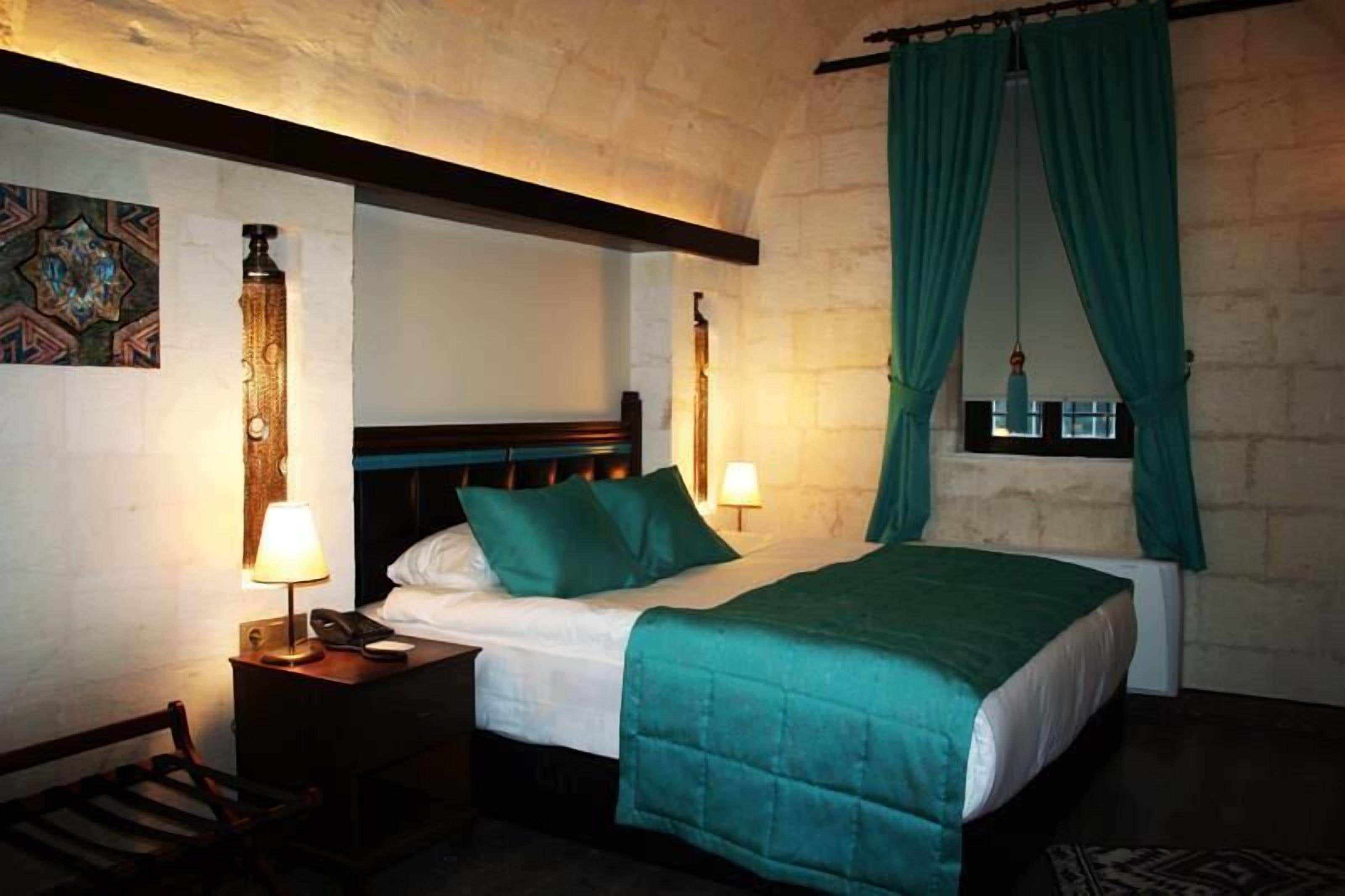 Quarto de hotel com cama, duas luminárias de cabeceira, mesa de cabeceira, uma cadeira e janela com cortinas.