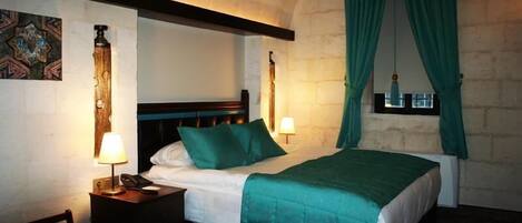 Quarto de hotel com cama, duas luminárias de cabeceira, mesa de cabeceira, uma cadeira e janela com cortinas.