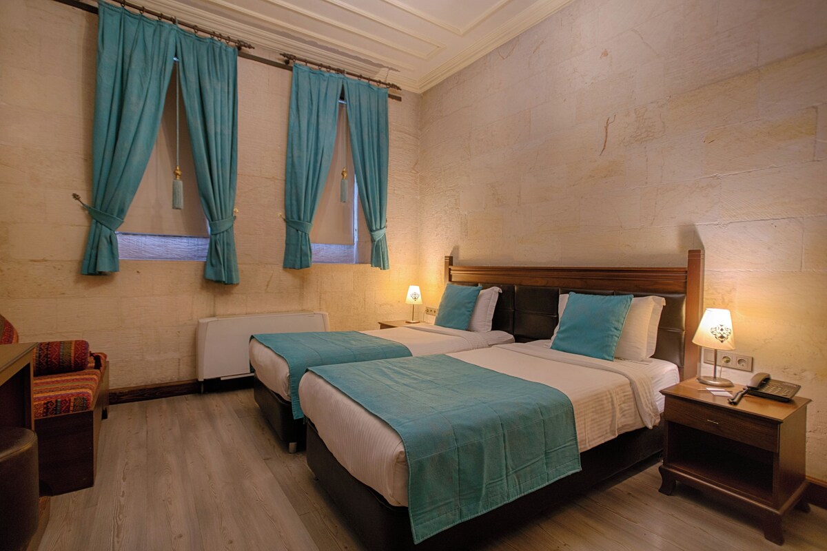 A hotel room with two beds, a nightstand with a lamp, a wooden headboard, and a window with curtains.