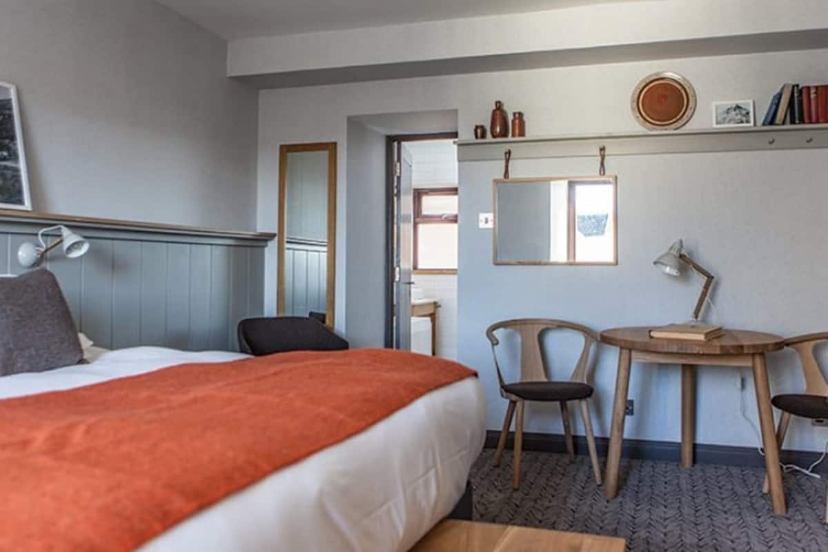 A hotel room with a bed, a desk, two chairs, a mirror, and a shelf with books and decorative items.