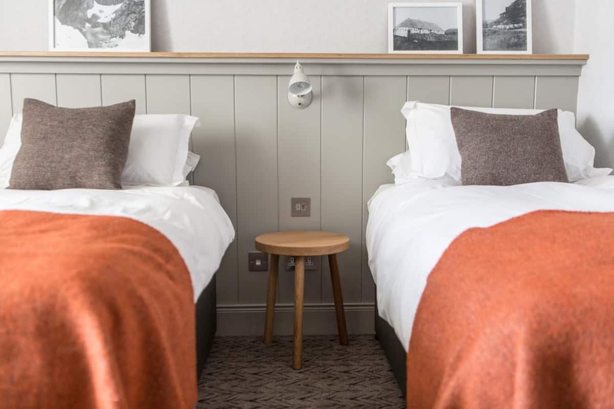 A hotel room with two beds, each with white bedding and brown pillows, a small wooden stool, and a wall-mounted light fixture.