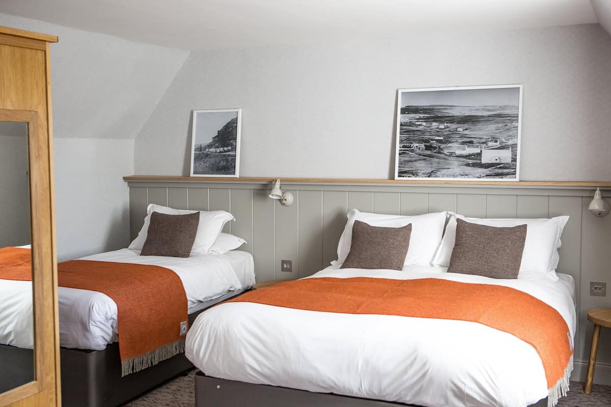 A hotel room with two beds, each with white bedding and orange throw blankets. There are two framed pictures on the wall above the beds.