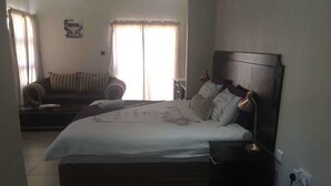 A bedroom with a large bed, a bedside table, a nightstand, a sofa, and a window with curtains. - Eagle Suites Guest House (Ghanzi)