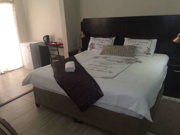A neatly made bed with a decorative pillow, a bedside table with a lamp, and a small refrigerator in a hotel room. - Eagle Suites Guest House (Ghanzi)