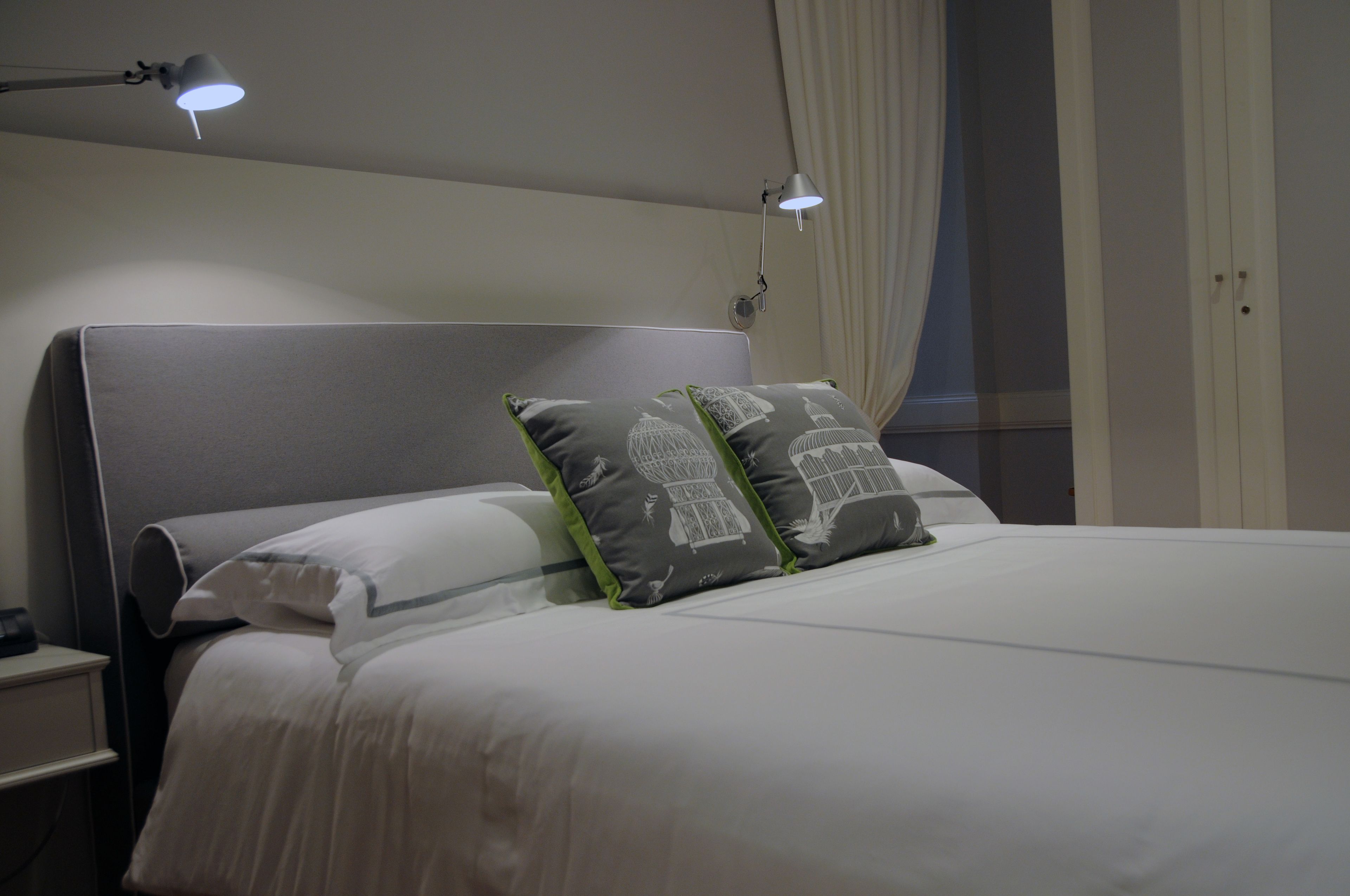 Una cama bien hecha con ropa de cama blanca y dos almohadas con estampado de pájaros. Hay una lámpara de noche en la mesita.