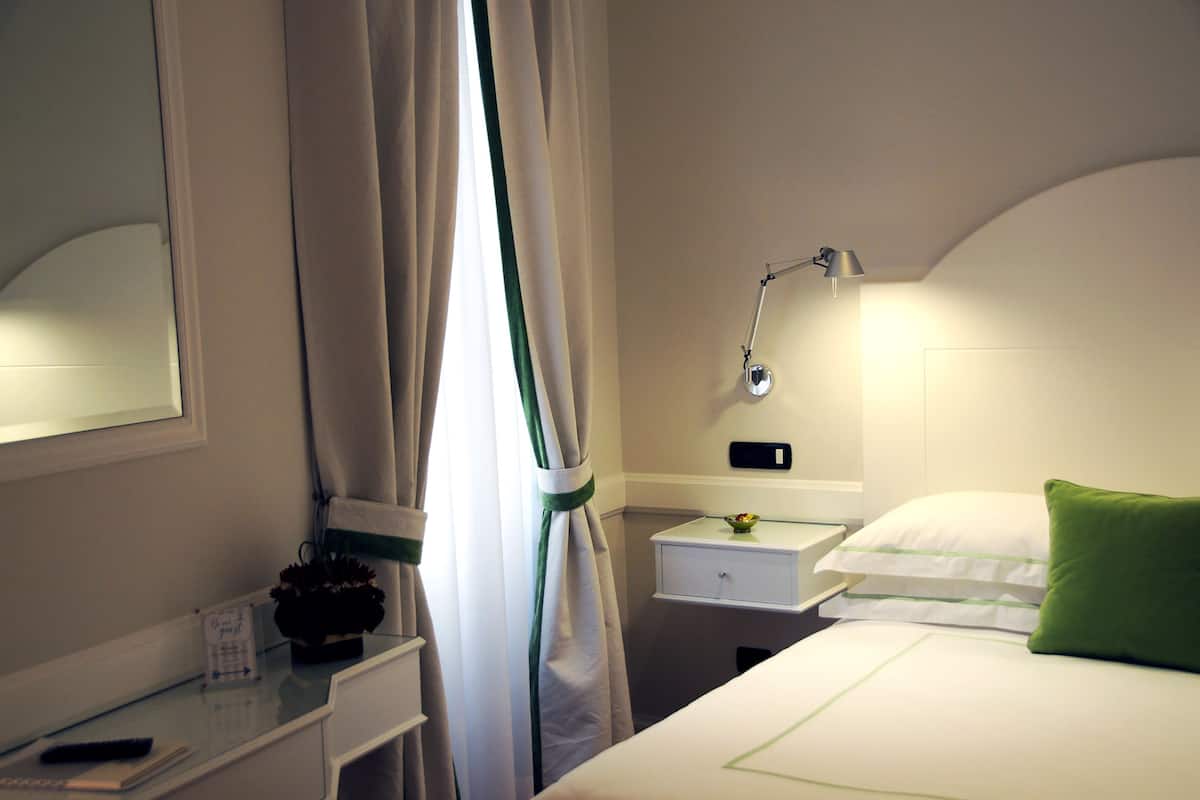 A hotel room with a bed, a nightstand, a lamp, a window with curtains, and a small table with a plant and a remote control.