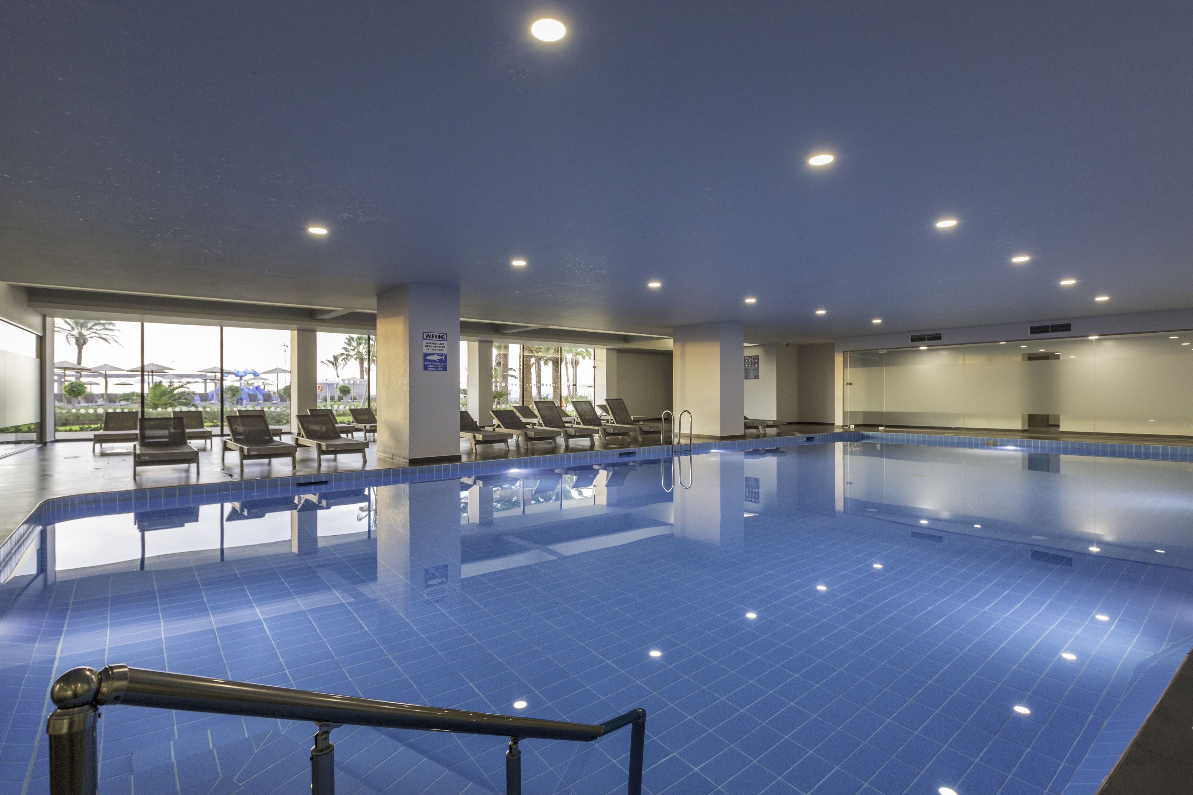 An indoor swimming pool with blue tiles, sun loungers, and a view of palm trees outside.