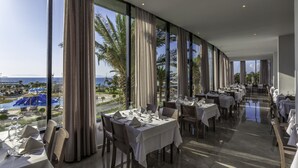 A restaurant with tables set for dining, large windows with curtains, and a view of the ocean and palm trees. - Akti Imperial Deluxe Resort & Spa Dolce by Wyndham - All inclusive (Rhodes)