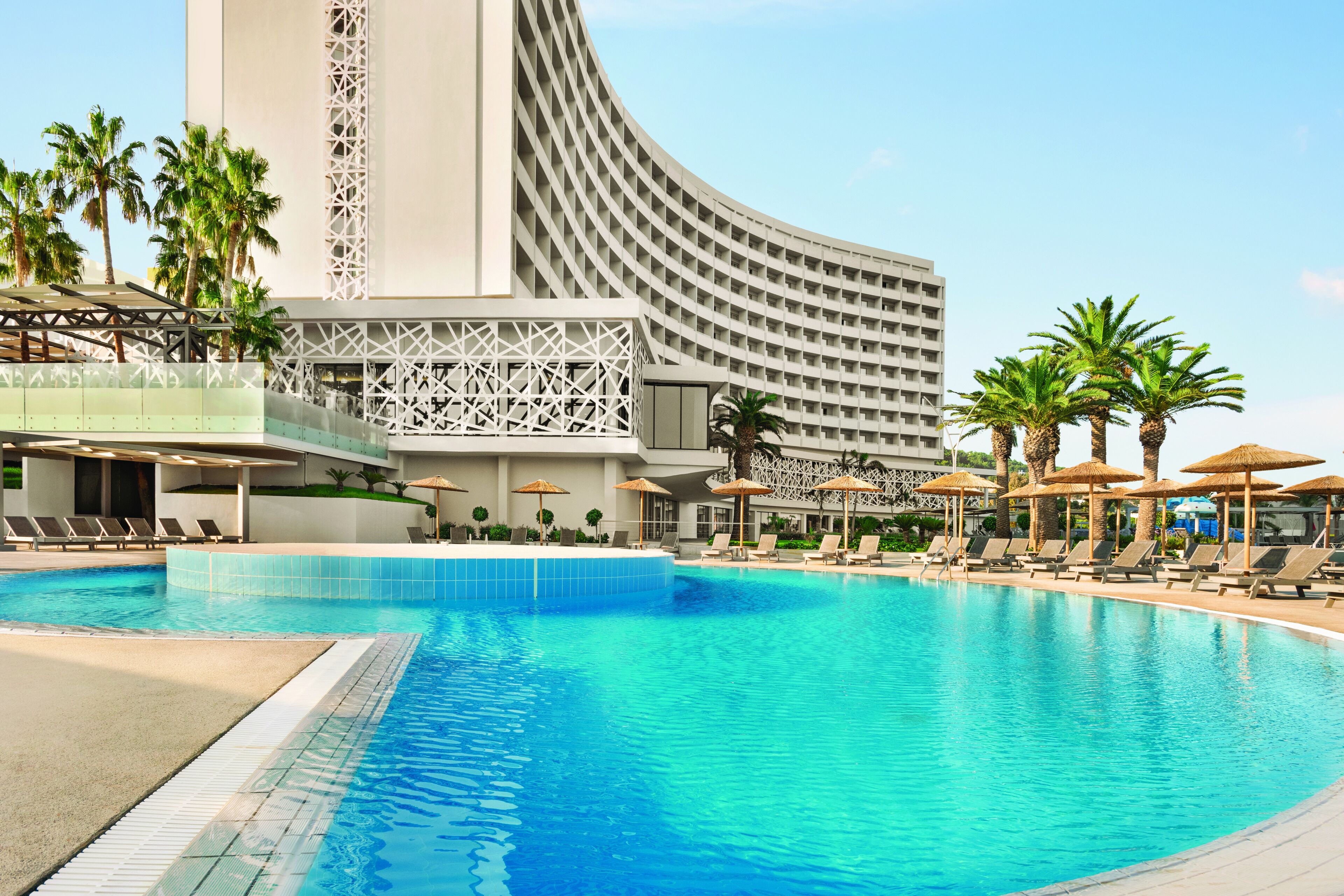 A swimming pool with lounge chairs and umbrellas, surrounded by palm trees and a modern building with a unique architectural design.