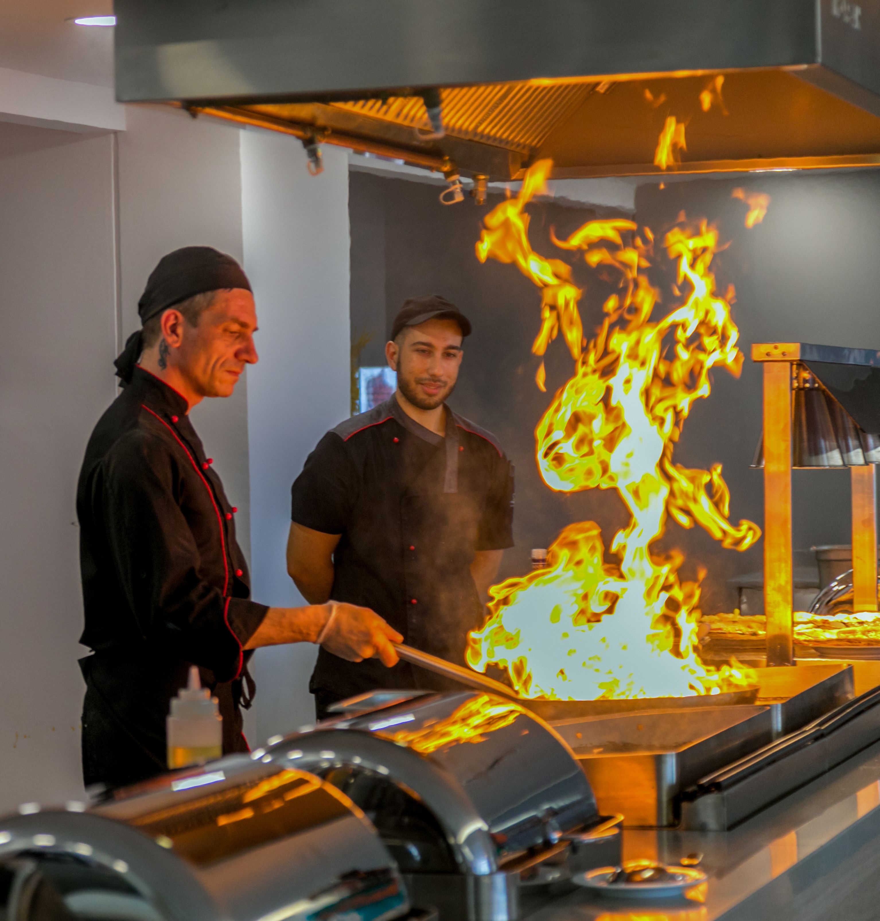Två kockar lagar mat över öppen eld på en grill i ett kök.