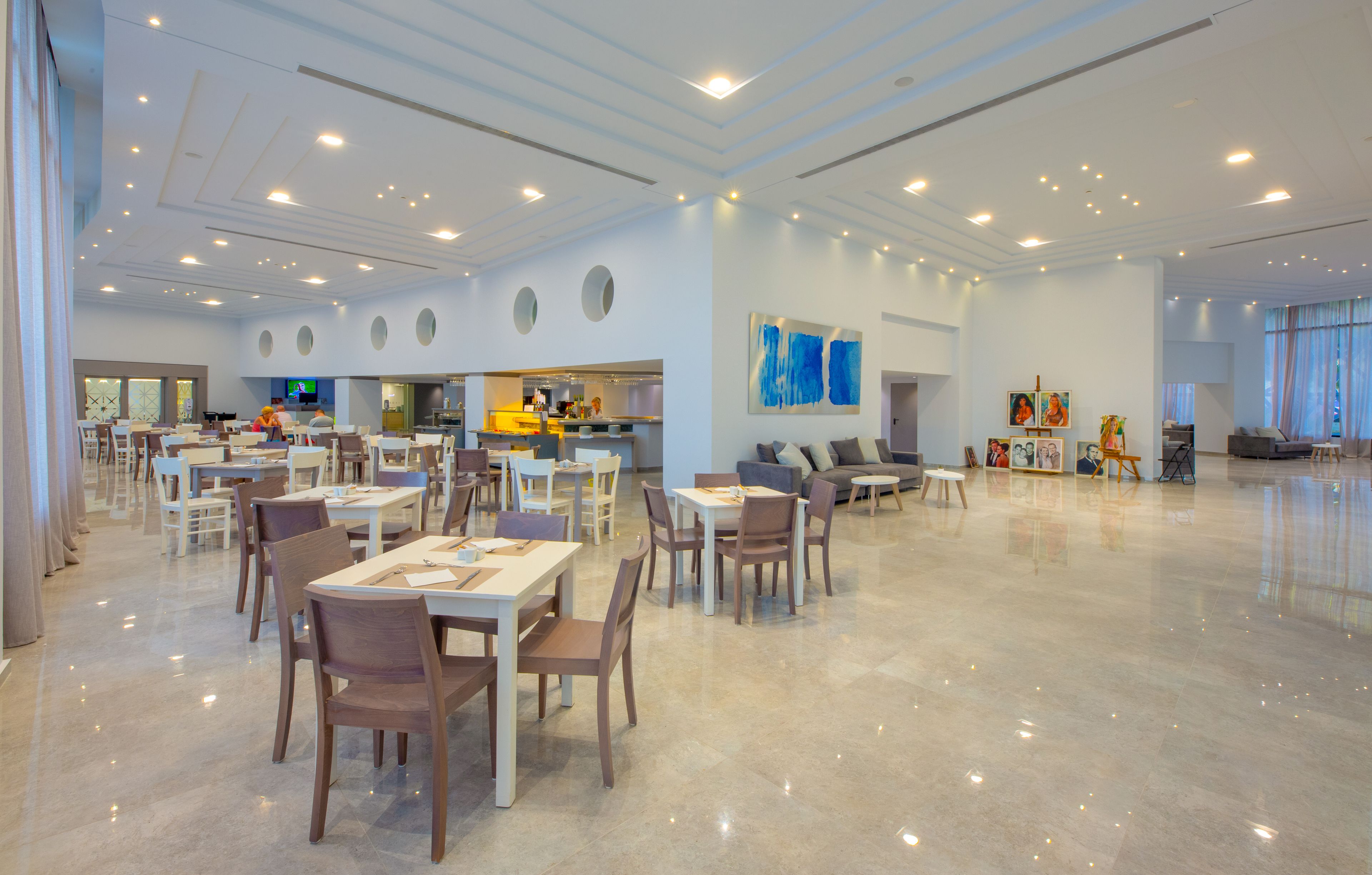A spacious dining area with multiple tables and chairs, a kitchen in the background, and a large abstract painting on the wall.
