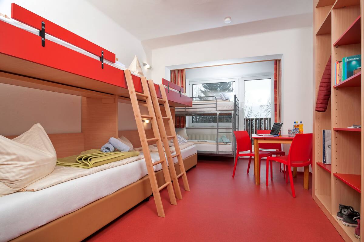 Una habitación tipo dormitorio con literas, un escritorio, sillas y una estantería.