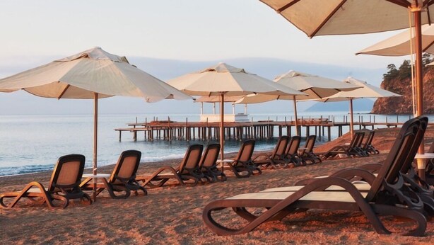 Een strandsetting met ligbedden en parasols, een houten pier die het water in reikt, en een kalme zee op de achtergrond.