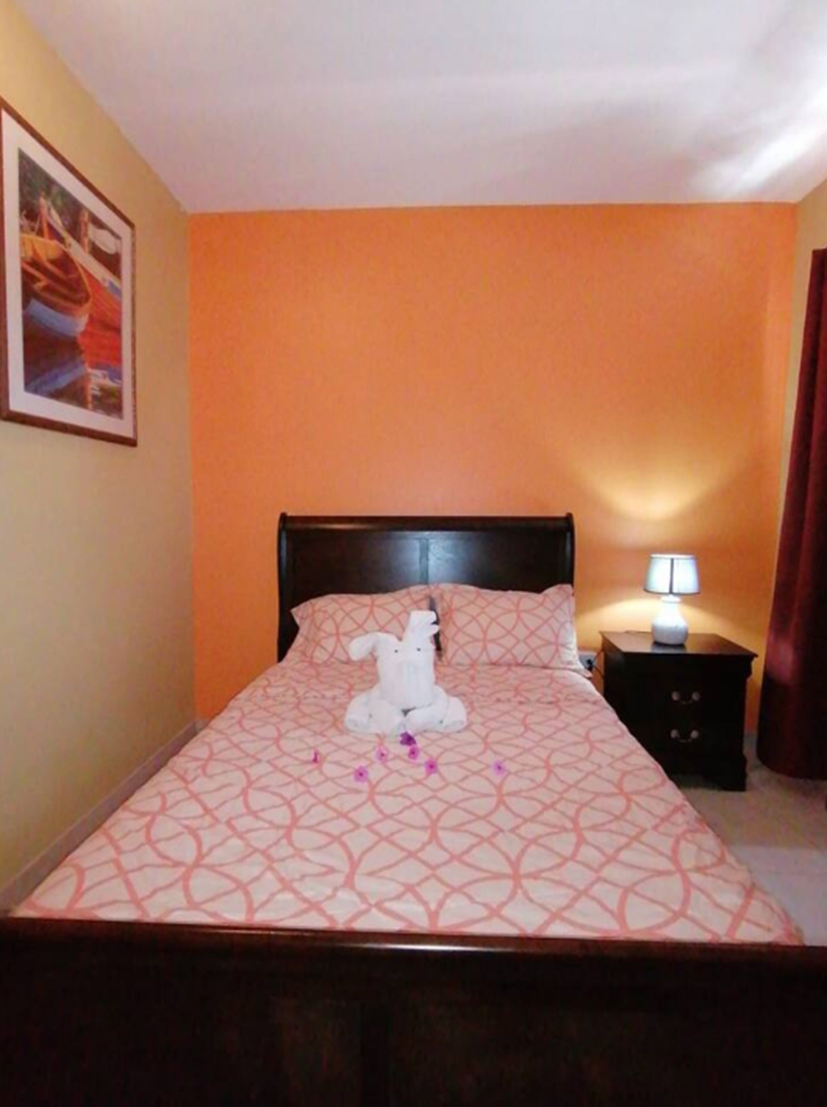 A neatly made bed with a patterned bedspread, a white towel shaped like a bunny, and a small nightstand with a lamp.