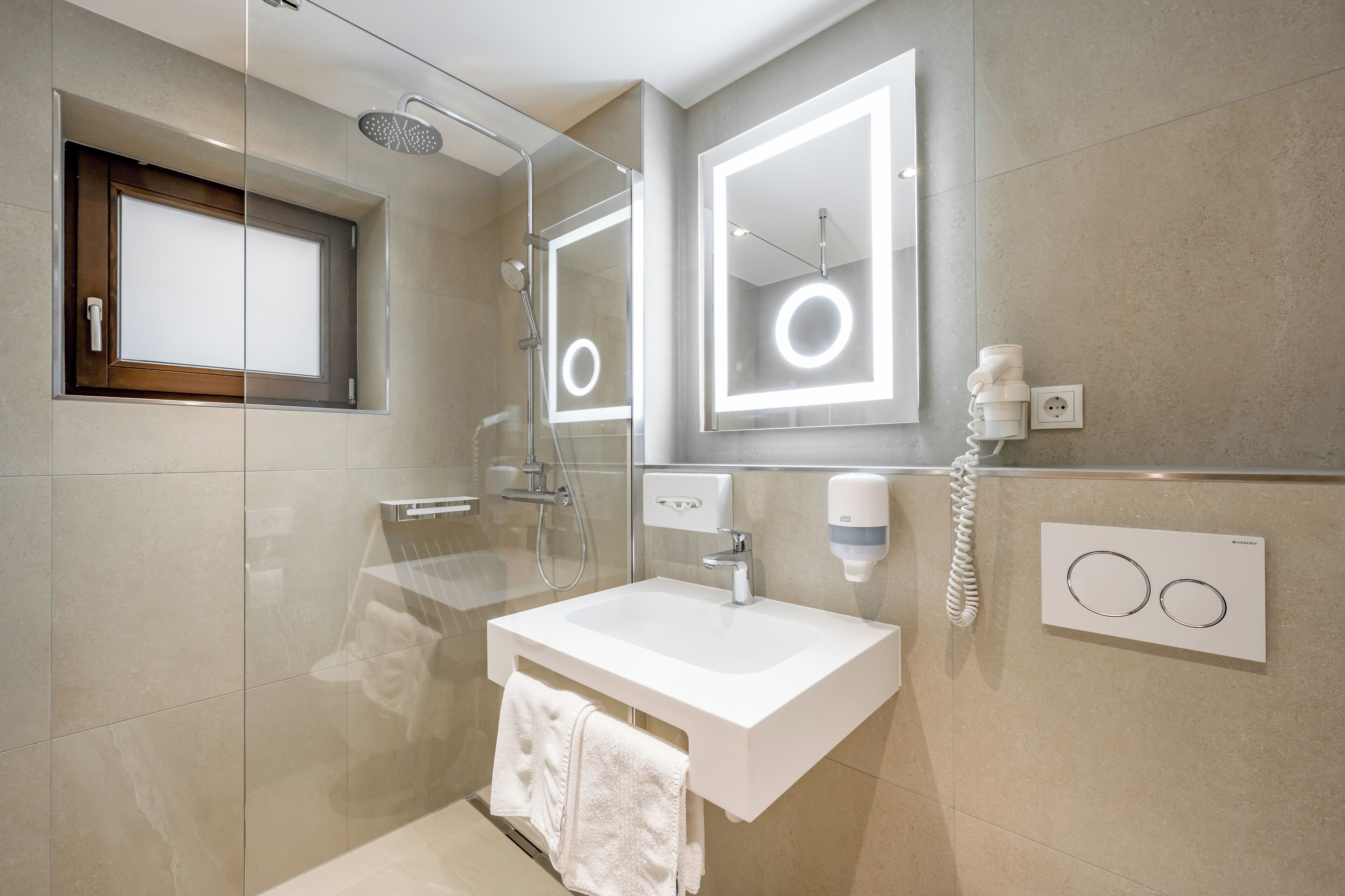 Un baño moderno con ducha envuelta en vidrio, un lavabo blanco y un espejo con iluminación circular.
