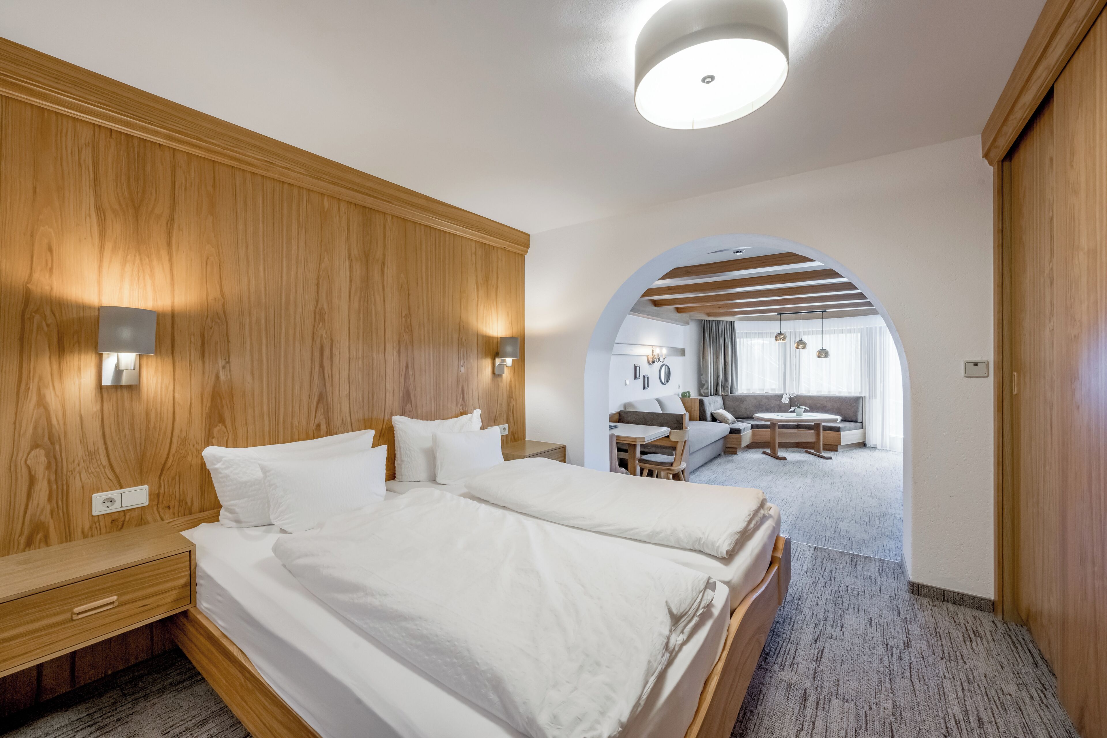 Una habitación de hotel moderna con cabecera de madera, una cama grande con ropa de cama blanca, un escritorio y una zona de estar con sofá y mesa.