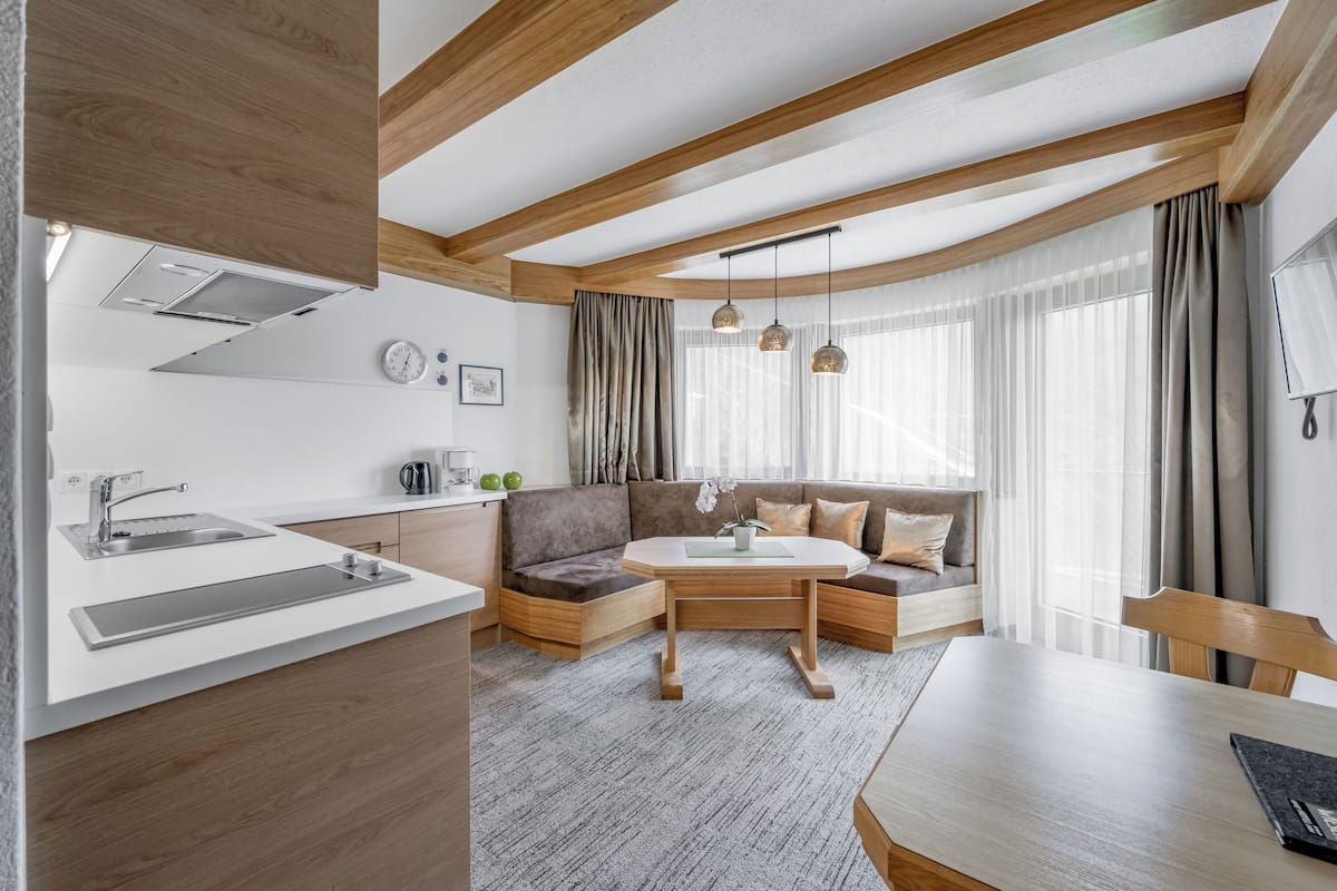 A modern kitchen with a built-in oven, a sink, and a countertop. There is a dining area with a table and chairs. The room has wooden beams on the ceiling and large windows with curtains.