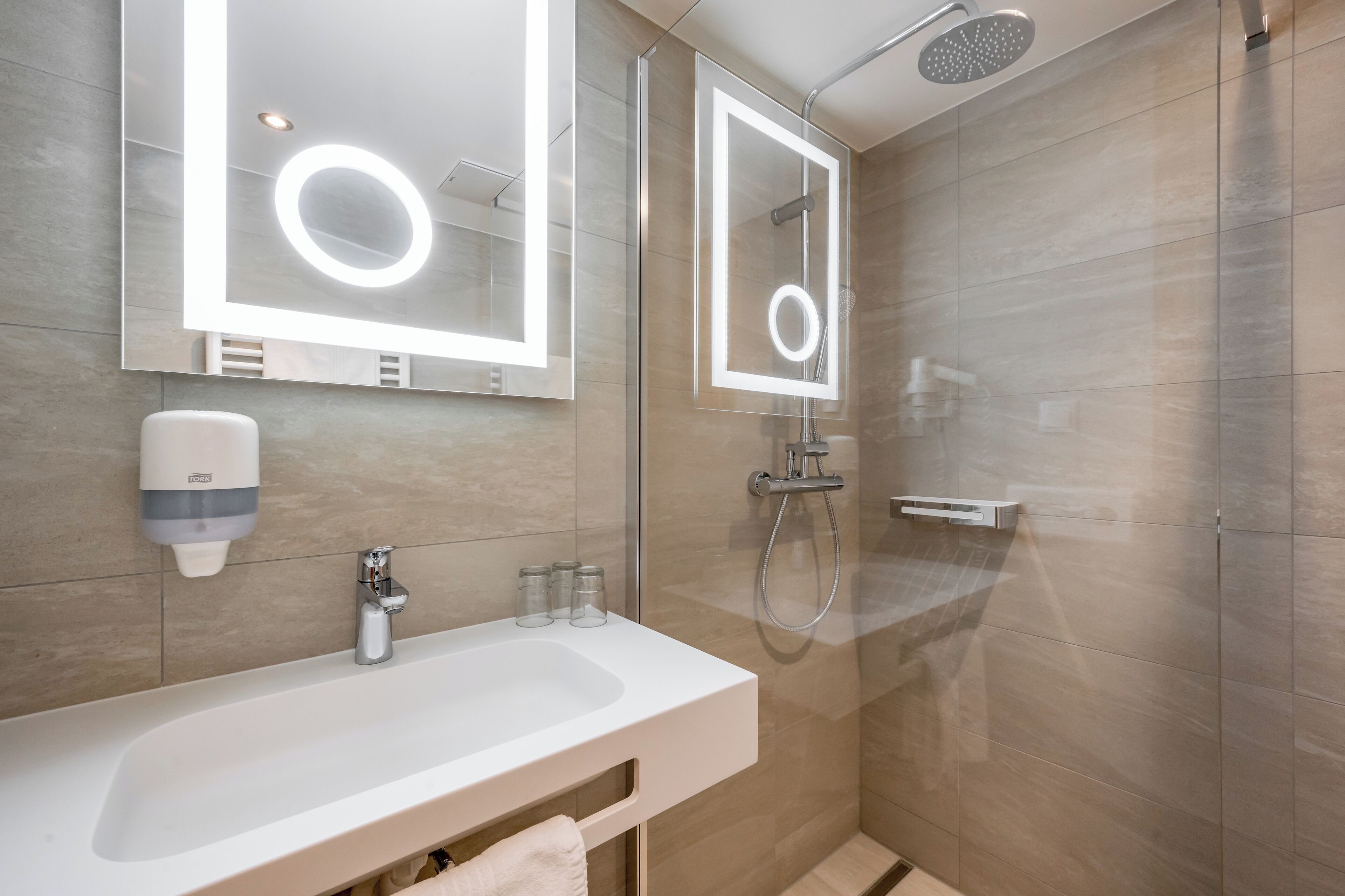 A modern bathroom with a large mirror, a sink, a handheld showerhead, and a soap dispenser.