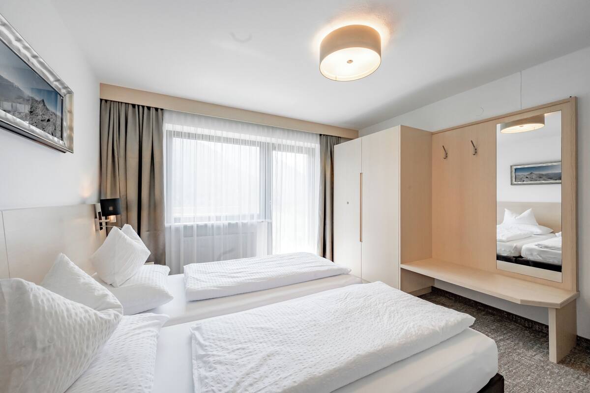A hotel room with two beds, a large mirror, a wardrobe, and a window with curtains.