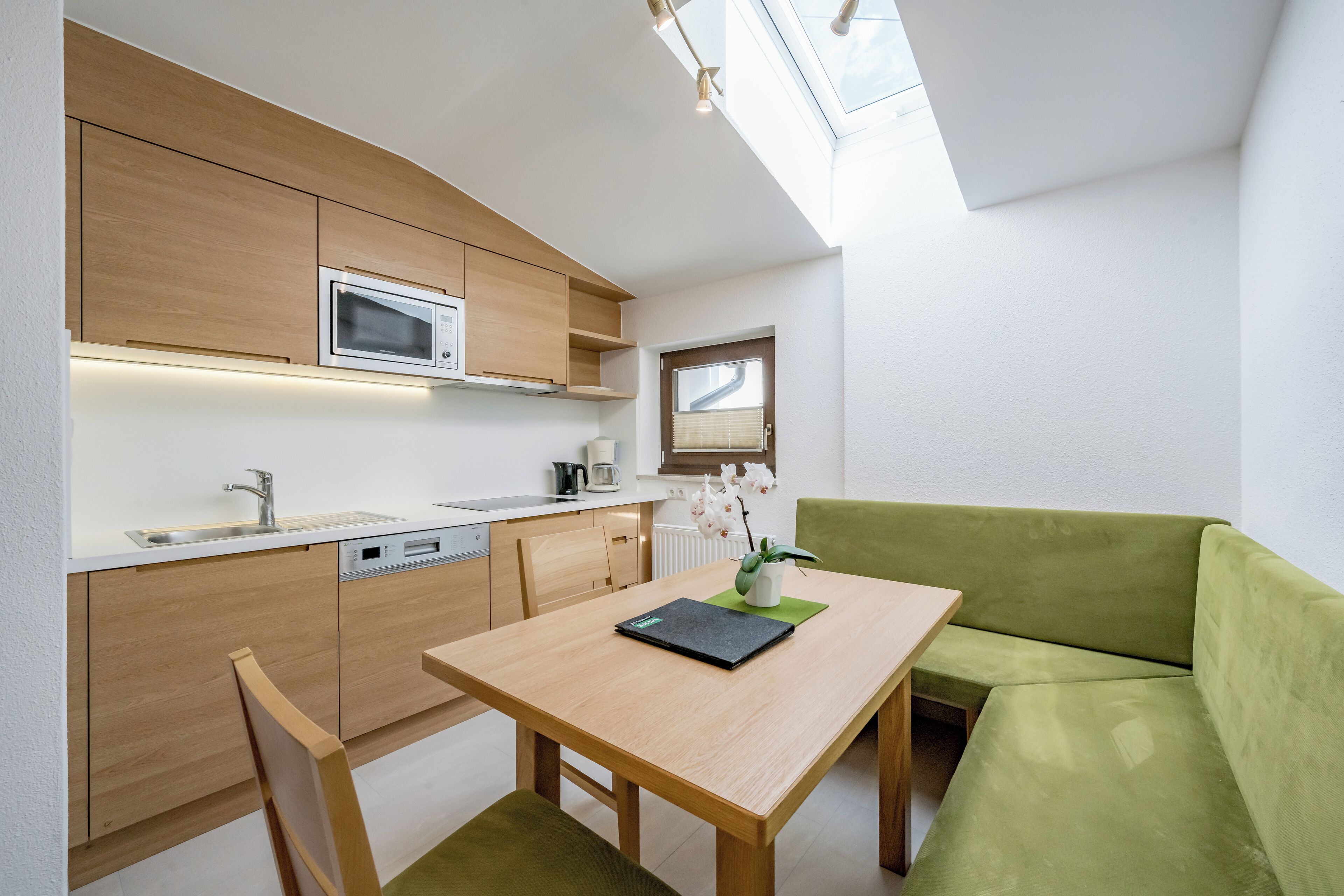 A compact kitchen with wooden cabinets, a microwave, and a sink. There's a dining table with a green cushion and a laptop. A green sofa is in the corner.