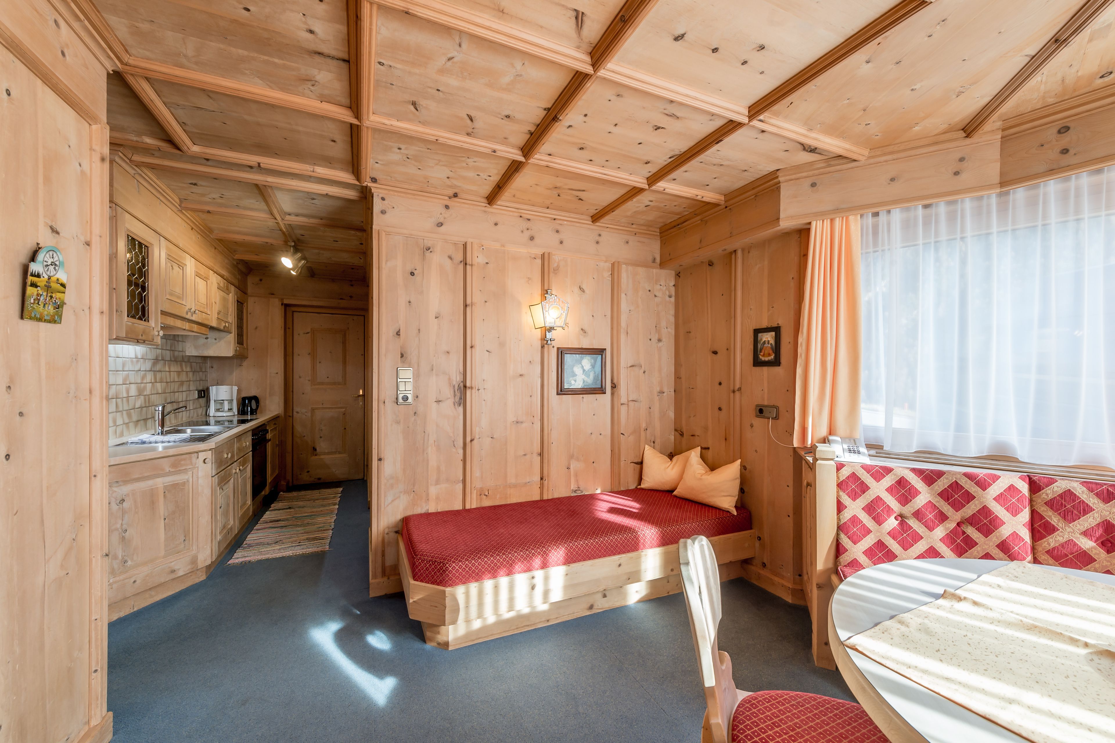 Interior de una cabaña de madera con cama, una kitchenette pequeña y una ventana con cortinas.