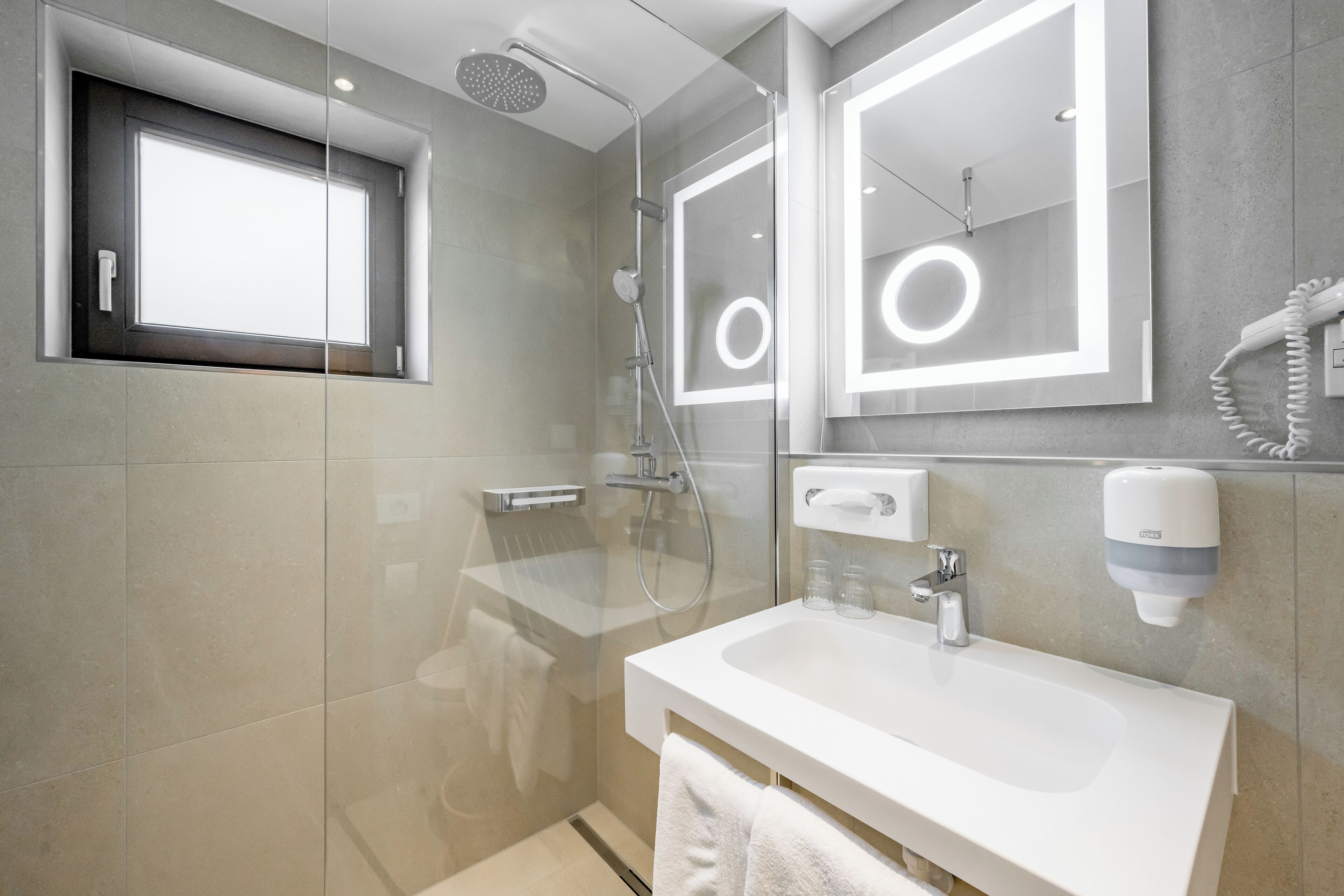 Un baño moderno con ducha envuelta en vidrio, un espejo grande con detalles de luz circulares, un lavamanos blanco y un perchero para toallas.