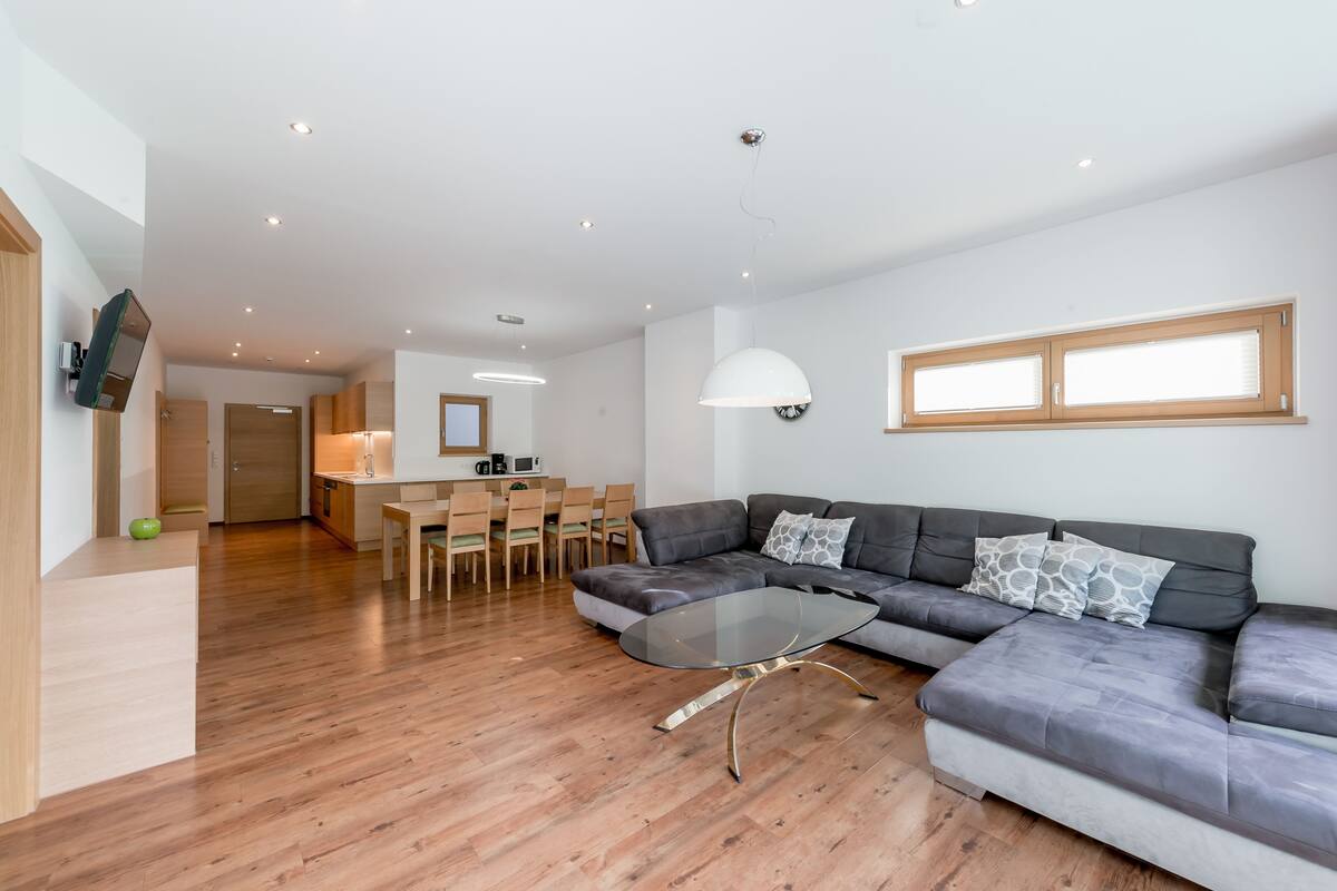 A modern living room with a sectional sofa, a glass coffee table, wooden flooring, a dining area with chairs, and a kitchen in the background.