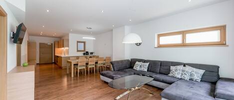 A modern living room with a sectional sofa, a glass coffee table, wooden flooring, a dining area with chairs, and a kitchen in the background.