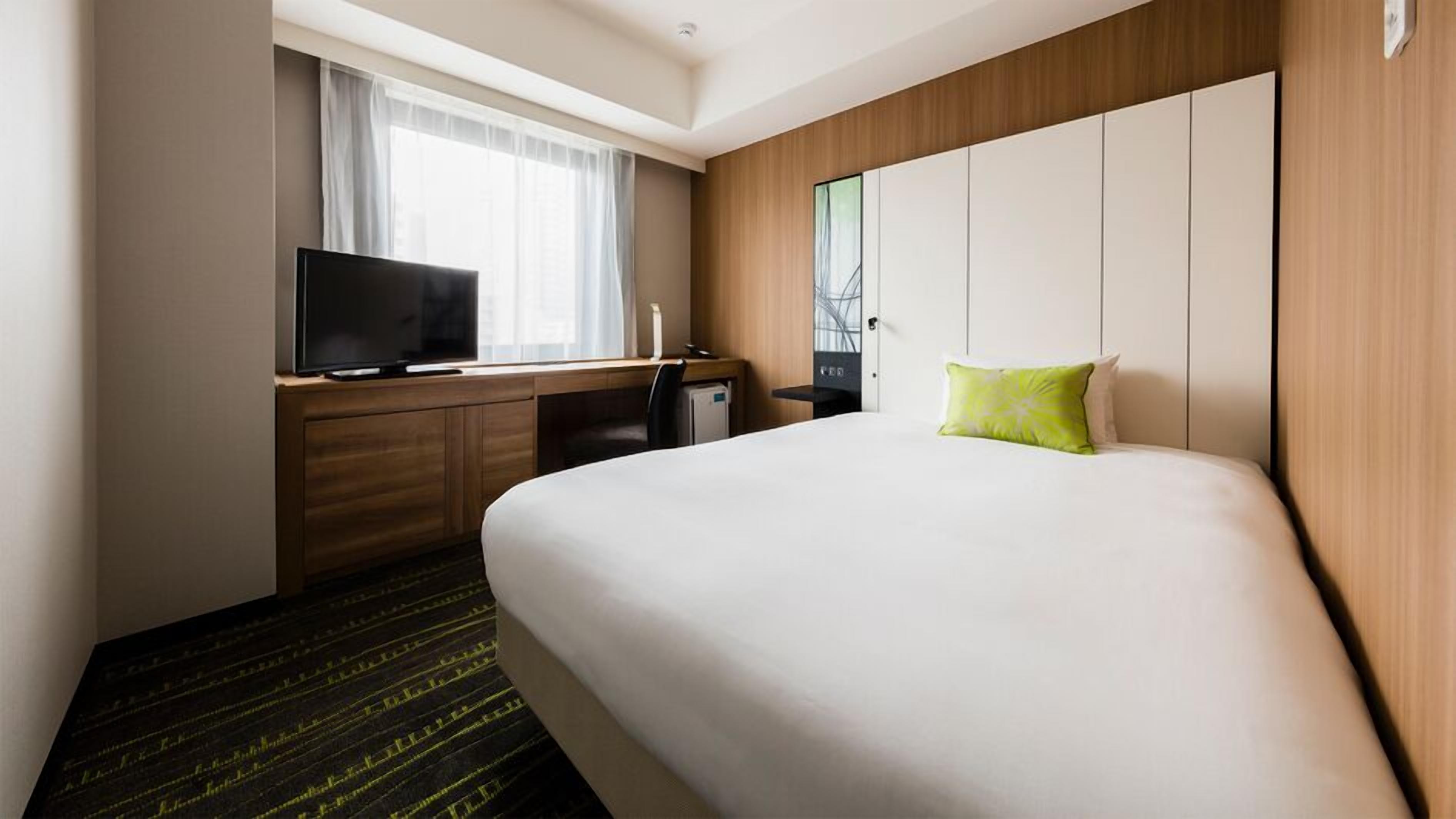 A hotel room with a bed, a desk with a TV, a small fridge, and a window with curtains.