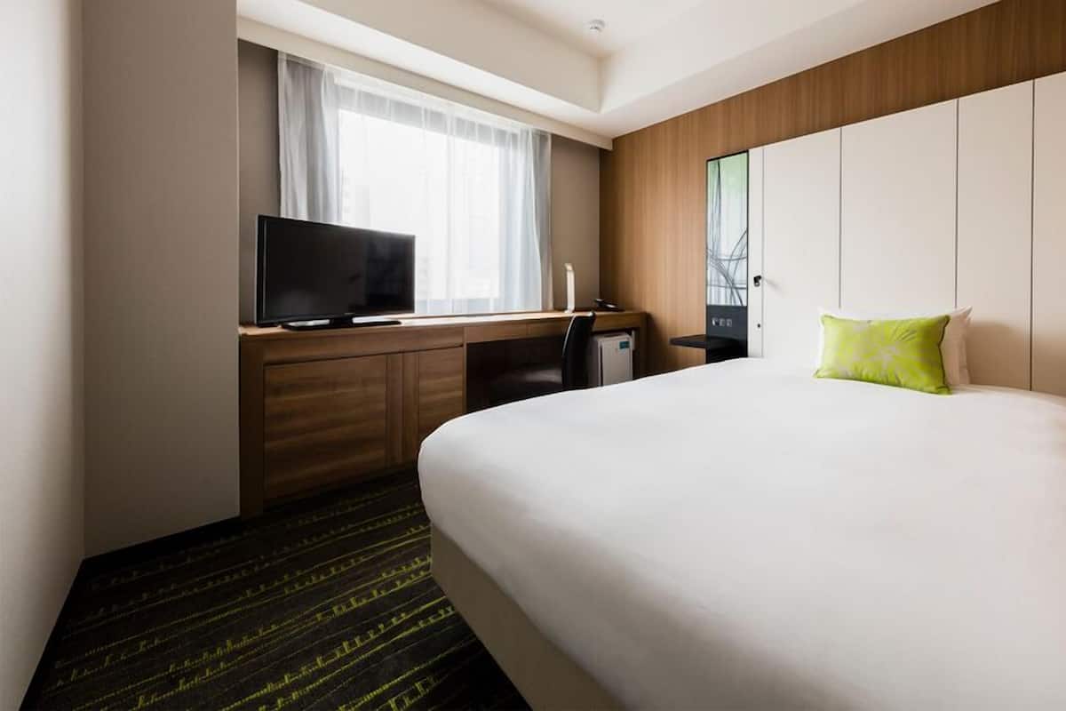 A hotel room with a bed, a desk with a TV, a small fridge, and a window with curtains.