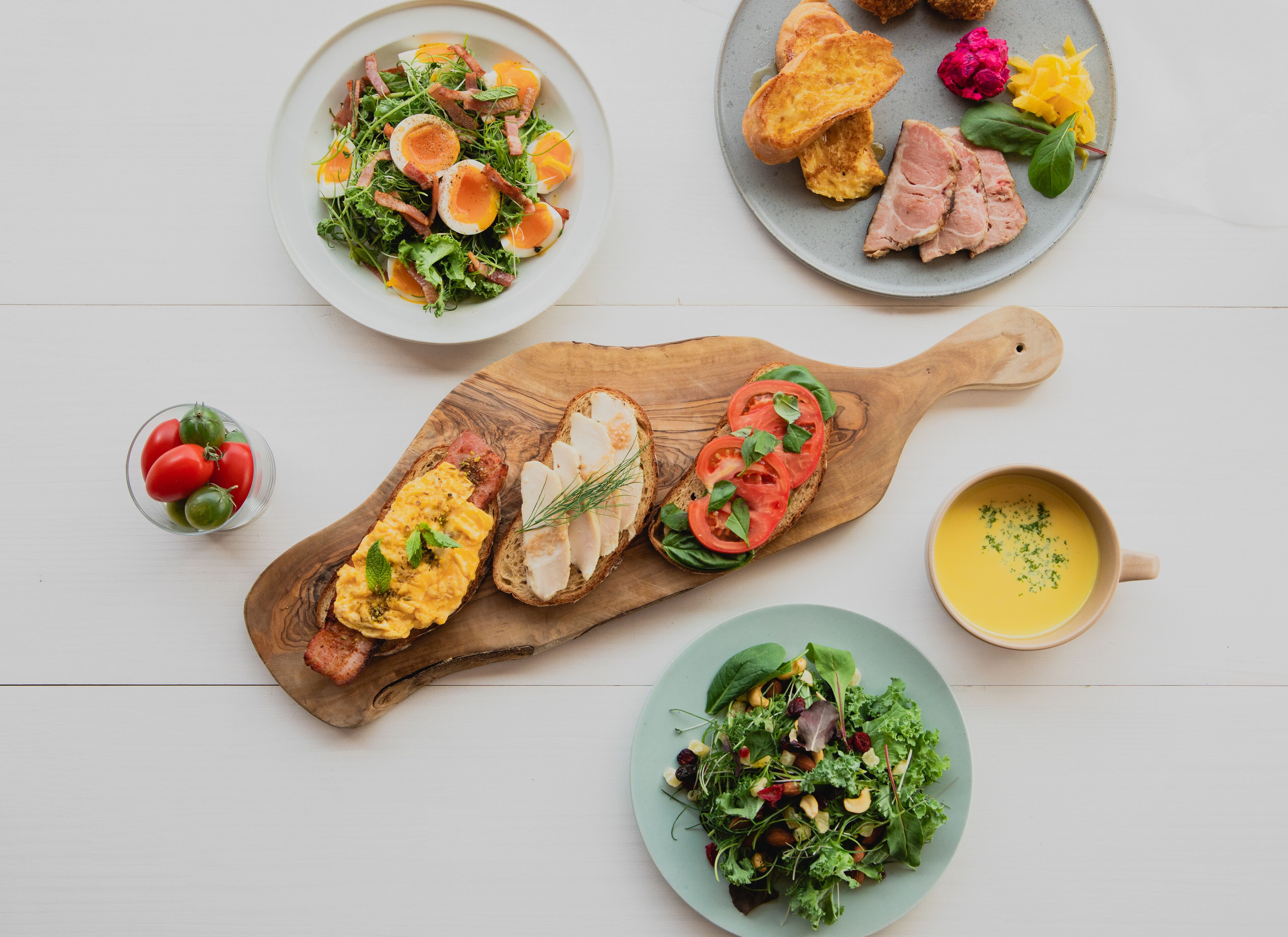 A variety of dishes including a salad, toast with eggs, and a bowl of soup, arranged on a wooden board and plates.