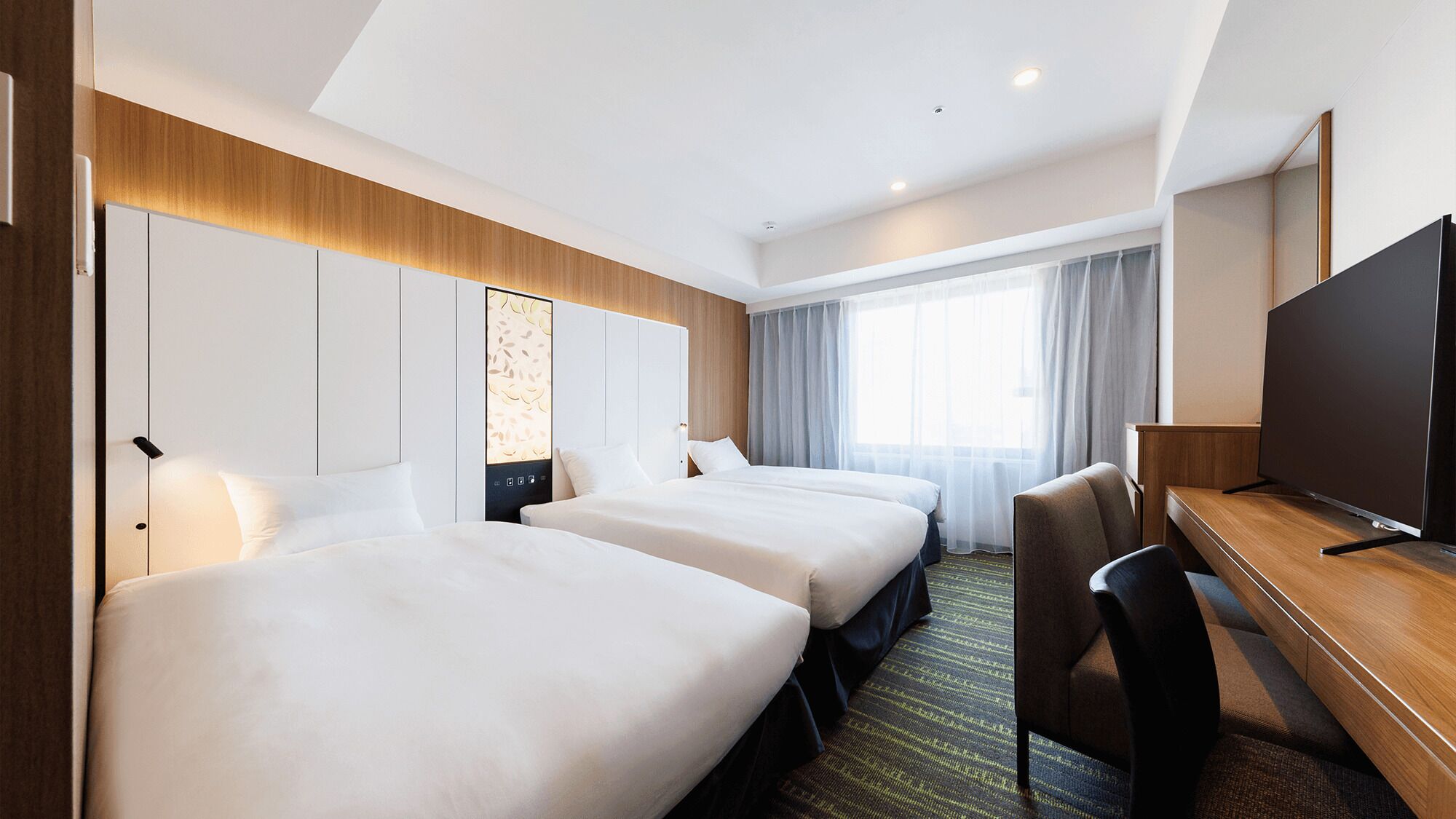 A hotel room with two beds, a desk, a TV, and a large window with curtains.