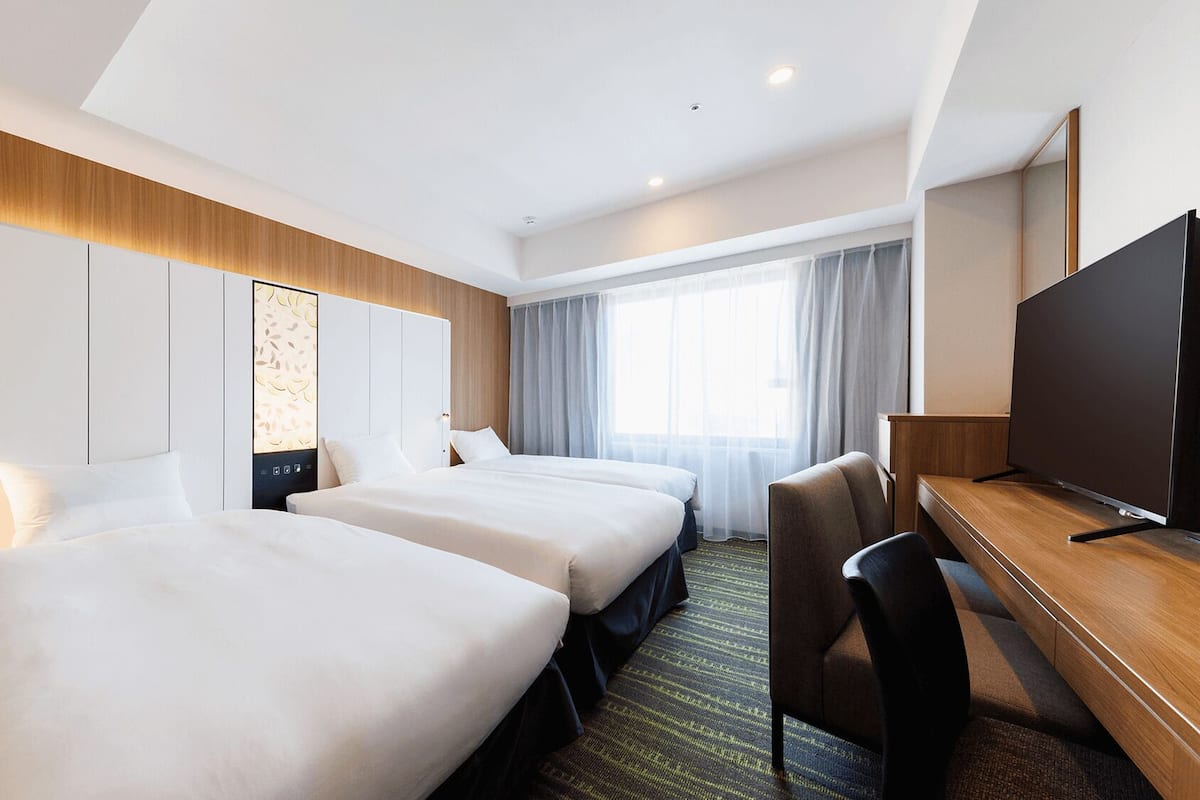 A hotel room with two beds, a desk, a TV, and a large window with curtains.