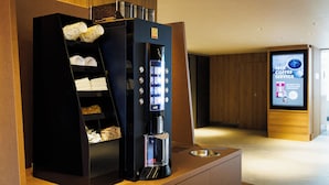 Une machine à café moderne dotée d’une interface numérique, d’une étagère pour les tasses et les serviettes, et d’un écran numérique affichant une publicité pour un service de café gratuit.