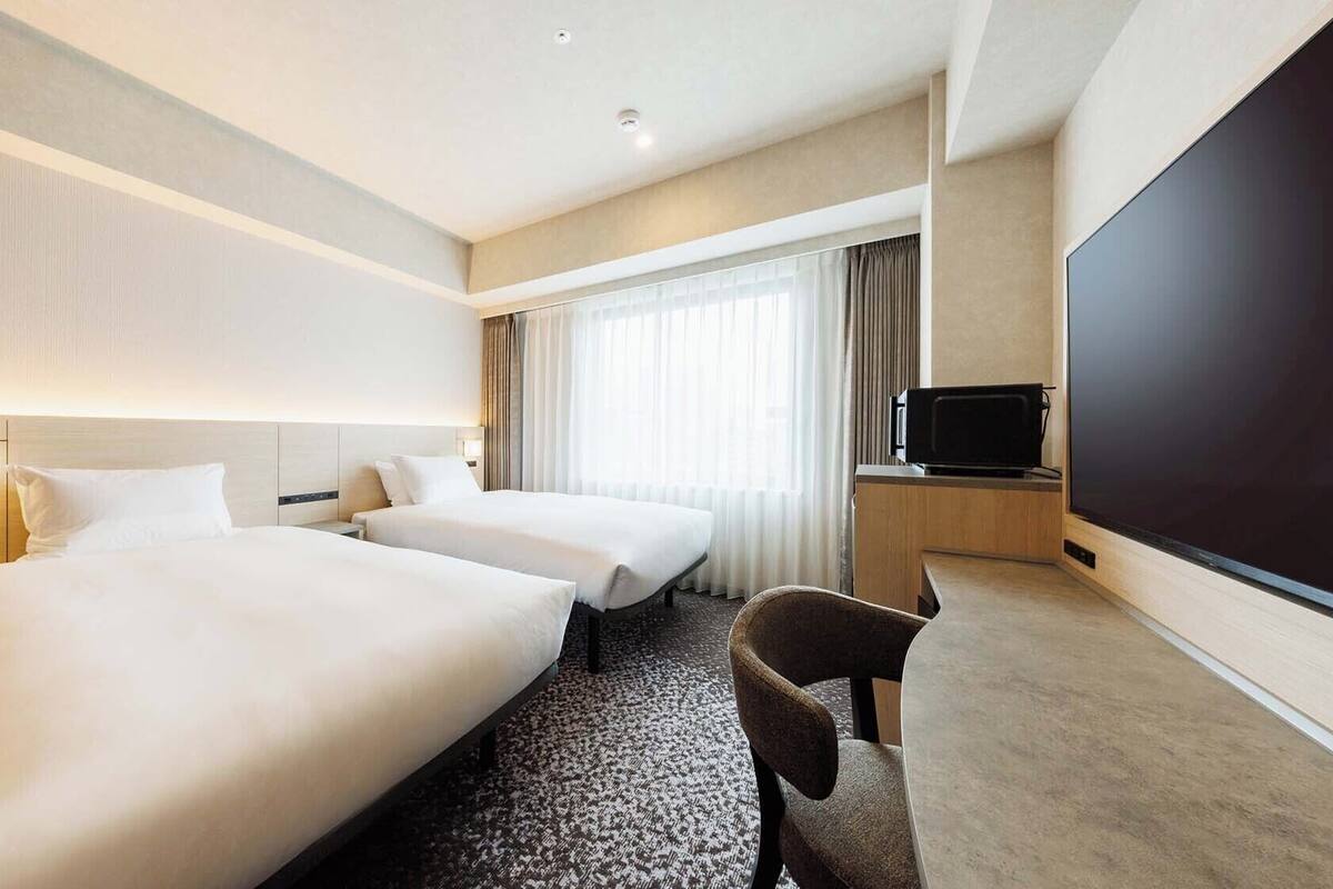 A hotel room with two beds, a desk with a flat-screen TV, and a large window with curtains.