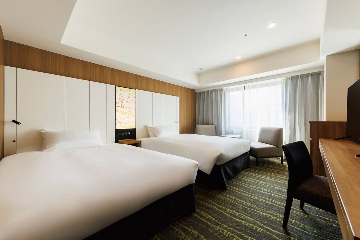 A hotel room with two beds, a desk, a TV, and a large window with curtains.