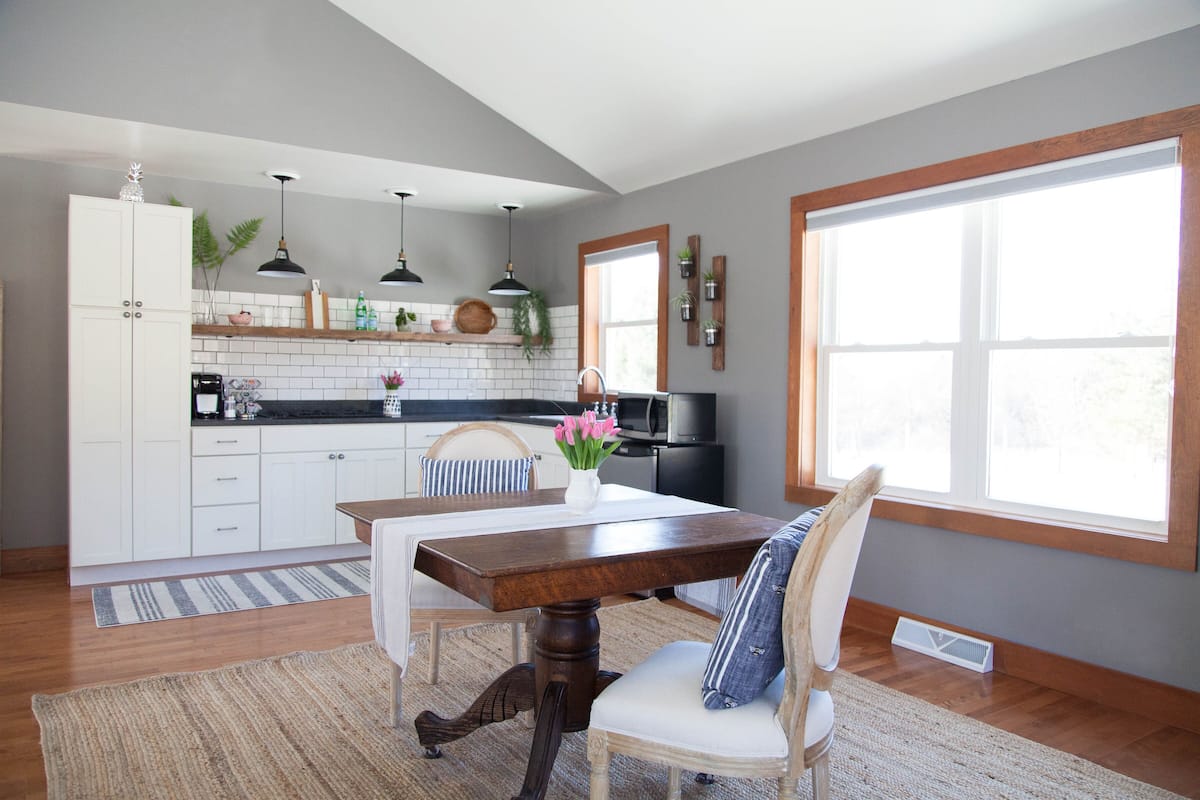A modern kitchen with white cabinets, a wooden dining table, and a large window.