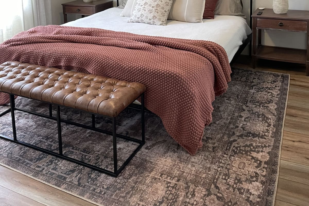 A bedroom with a bed, a bench, a ceiling fan, a nightstand, a lamp, and a painting on the wall.