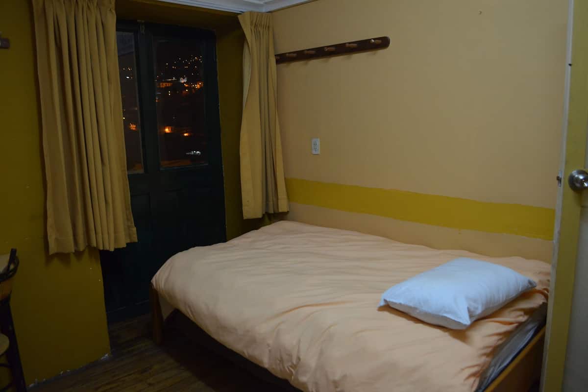 Una habitación pequeña con cama individual, cabecera de madera, ventana con cortinas y vistas a luces de la ciudad.