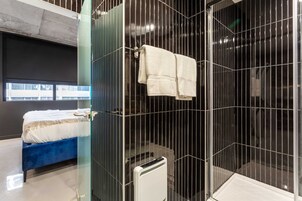 Une salle de bain moderne avec une douche à l’italienne en verre, une serviette blanche accrochée à la porte, et un lit bleu dans la pièce adjacente.