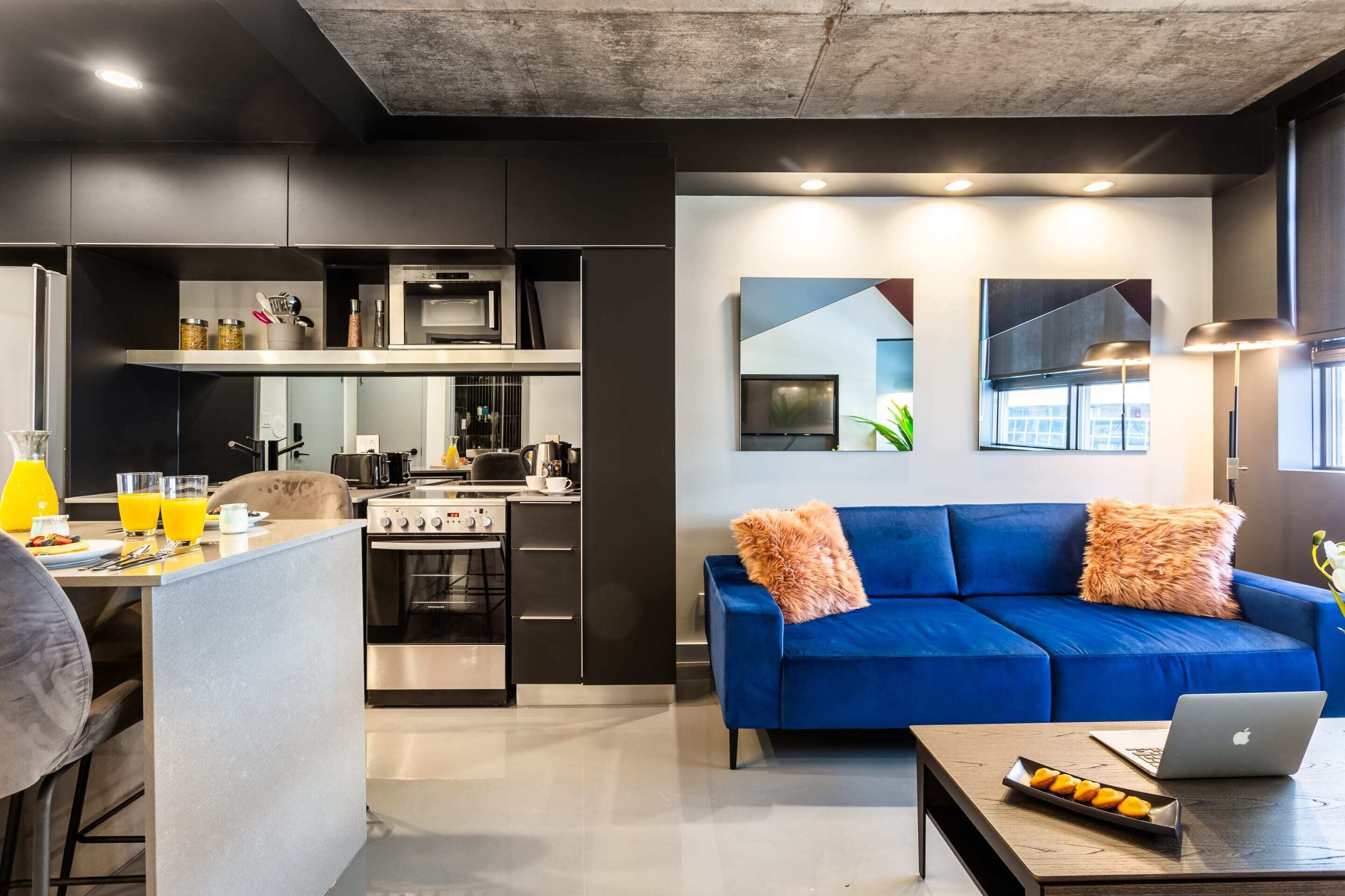 A modern kitchen with a built-in oven and microwave, a dining area with bar stools, and a living room with a blue sofa and a laptop on a coffee table.