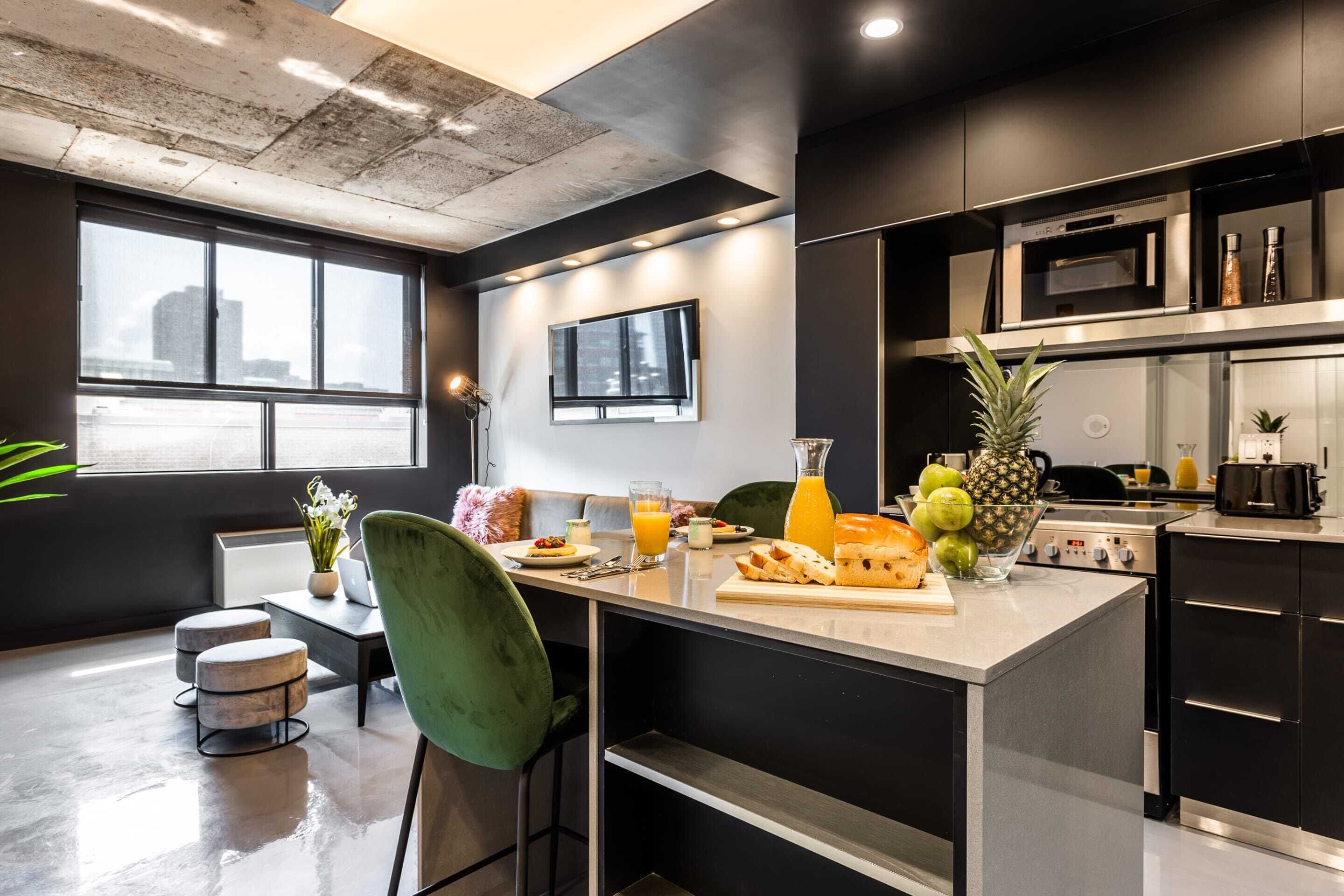 Una cocina moderna con una isla central, un área de comedor con una silla verde y un ventanal con vistas a la ciudad.