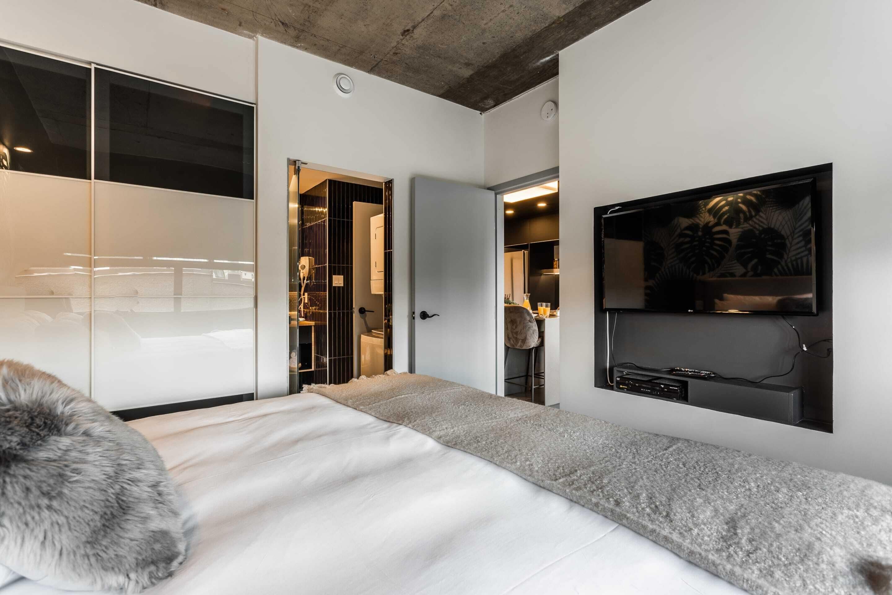 A modern bedroom with a large bed, a TV mounted on the wall, and a walk-in closet.